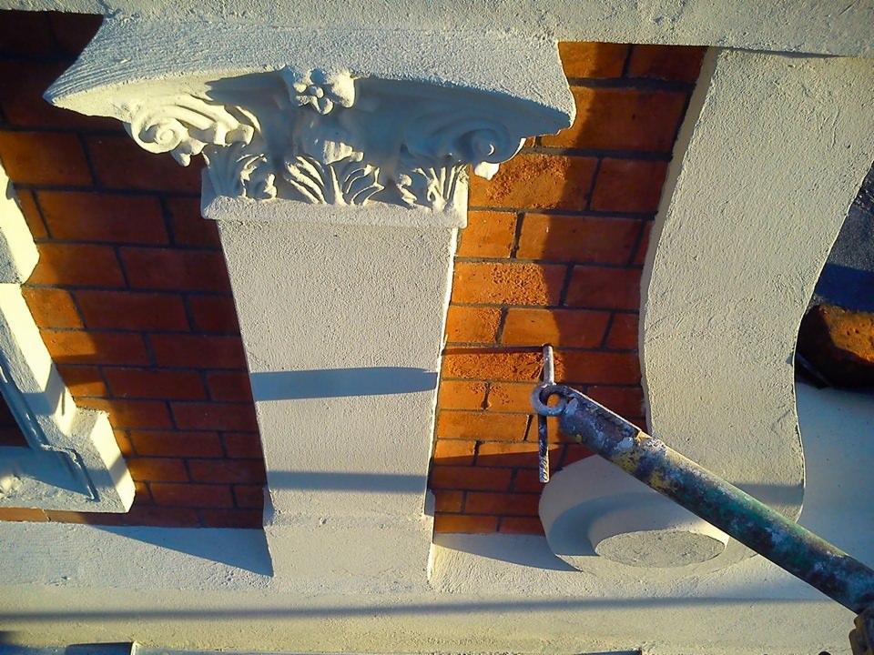 Renowacja detali architektonicznych: fragment kolumny z kapitelem zdobionym motywami roślinnymi, ceglana ściana w tle i fragment rusztowania z metalowym hakiem.