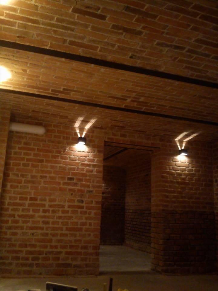 Wnętrze z ceglanym sklepieniem łukowym, oświetlone kinkietami, z widoczną konstrukcją nośną