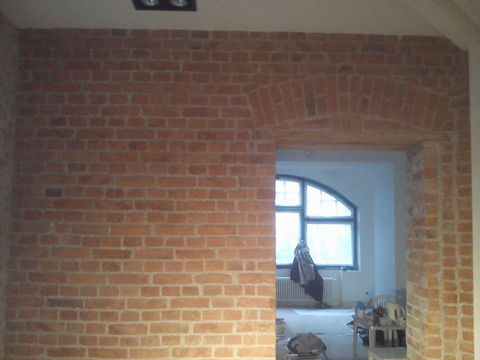Ceglana ściana z łukowym przejściem, widoczne prace remontowe w pomieszczeniu obok, okno z łukiem w tle.