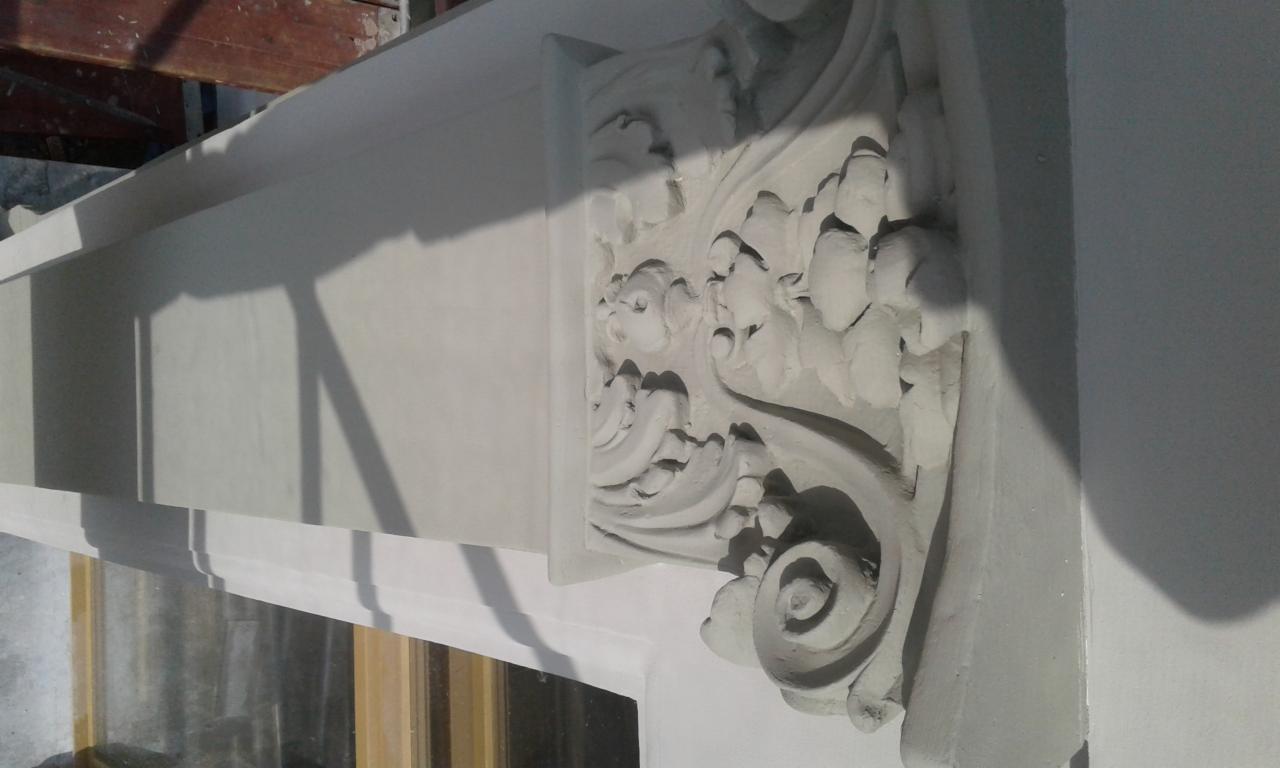 Detal dekoracyjny w postaci płaskorzeźby z motywami roślinnymi, prawdopodobnie wykonany z gipsu lub stiuku, umieszczony na białej ścianie w pobliżu okna.