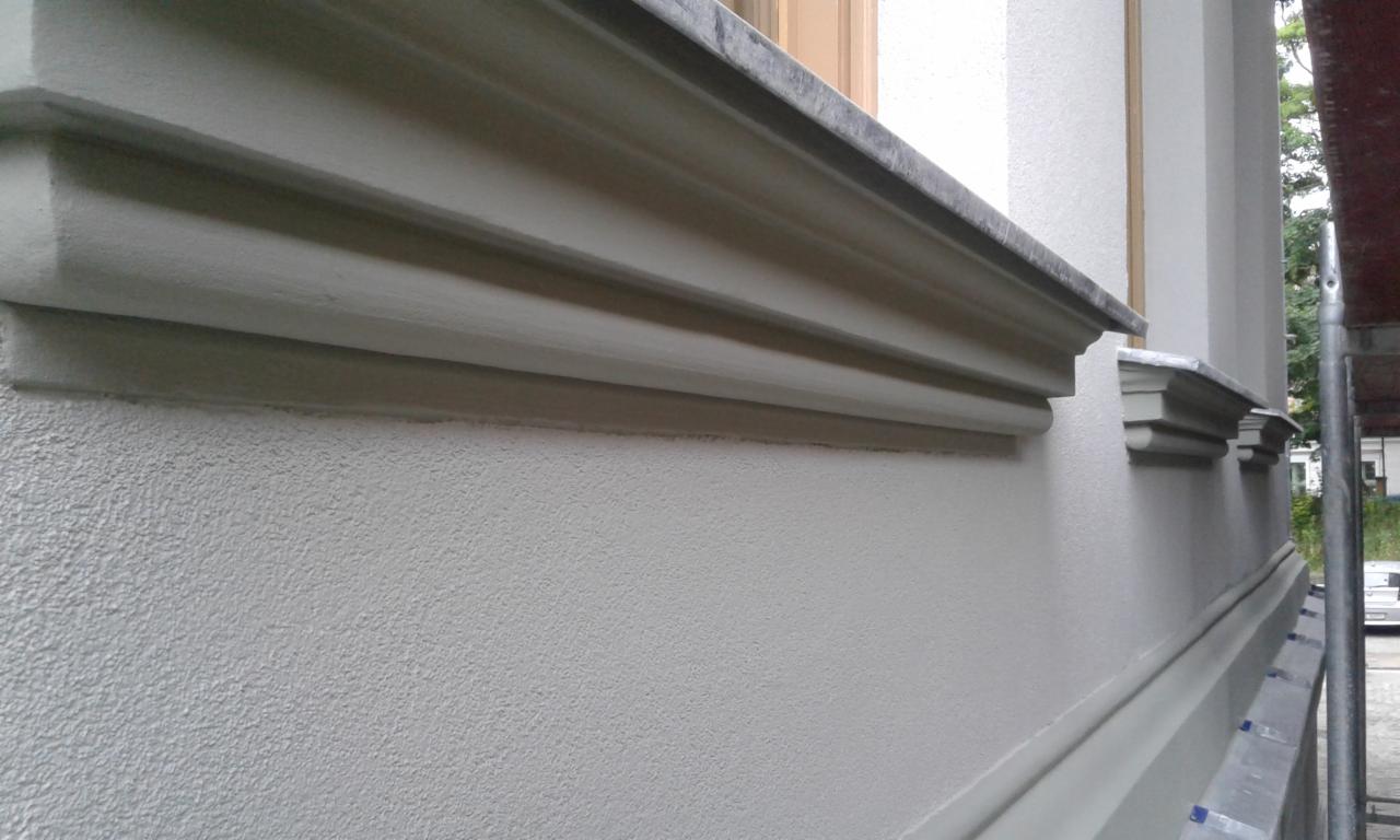 Odnowiona elewacja budynku z dekoracyjnymi elementami architektonicznymi, widoczne sztukaterie okienne i gzymsy, świeżo pomalowane na jasny kolor, z fragmentem rusztowania po prawej stronie.