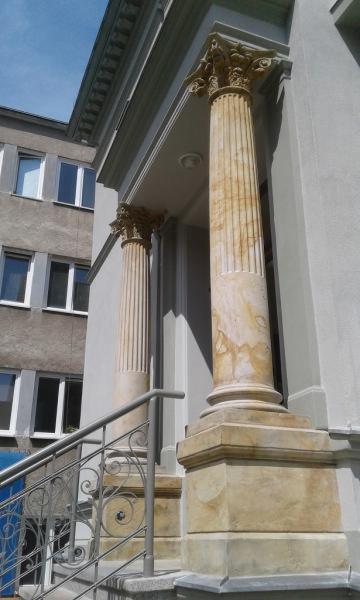 Odrestaurowane kolumny z kapitelami przed wejściem do budynku, widoczne ślady renowacji na kamieniu, metalowa balustrada schodów z ozdobnymi elementami.