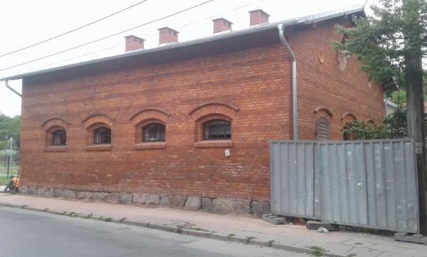 Dwukondygnacyjny budynek z czerwonej cegły z łukowymi oknami, widoczne cztery kominy na dachu, fragment chodnika i metalowe ogrodzenie z boku.