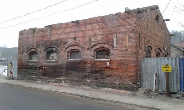 Zniszczony, ceglany budynek z łukowymi oknami, częściowo zabezpieczony metalowym ogrodzeniem z żółtą tabliczką ostrzegawczą, widoczny z poziomu ulicy.