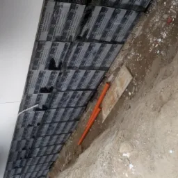 Warstwy papy termozgrzewalnej z widocznymi napisami 'Foundation 4.0 Speed Profil SBS' ułożone na dachu, obok wykop z pomarańczowymi rurami i ziemią.