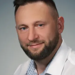 Portret uśmiechniętego mężczyzny z brodą w białej koszuli z wzorzystym kołnierzykiem na szarym tle.