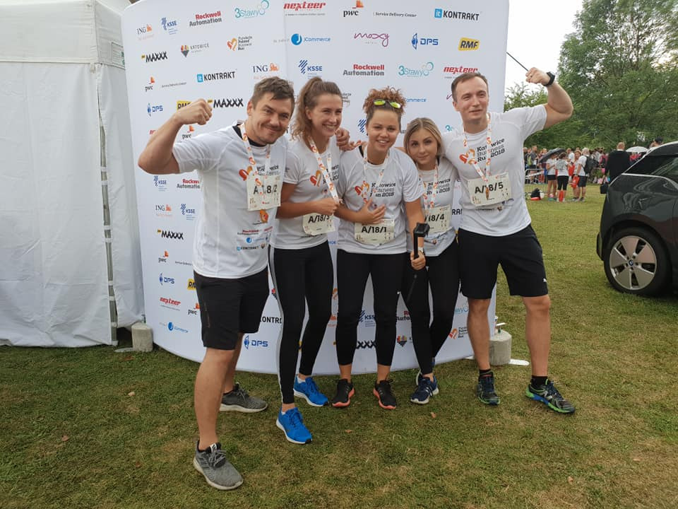 Grupa pięciu uśmiechniętych osób w sportowych strojach, prezentujących muskuły na tle ścianki sponsorskiej po biegu, na tle zielonej trawy i szarego samochodu.