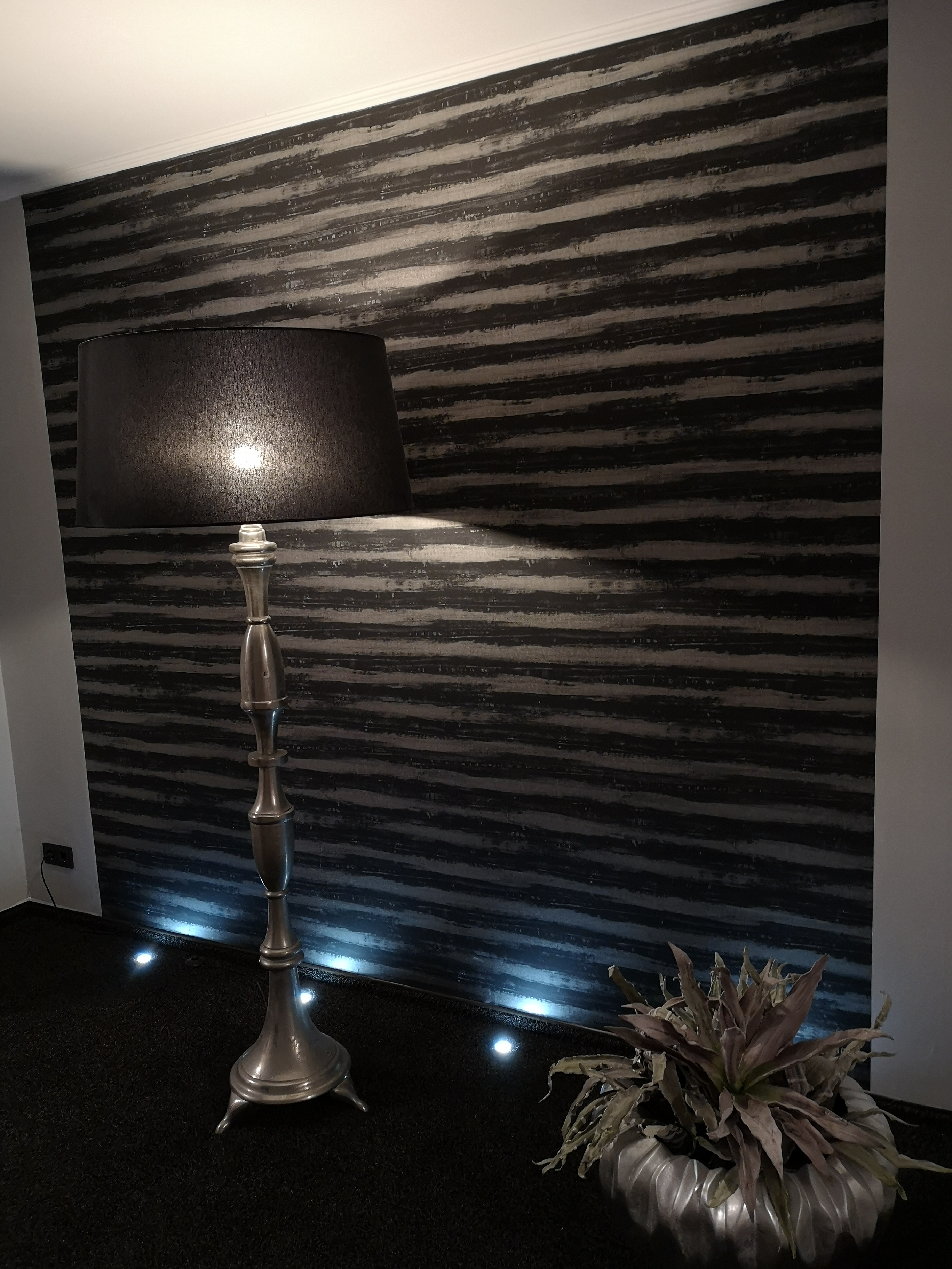 Eleganckie wnętrze z lampą podłogową w stylu art deco i dekoracyjną ścianą w poziome pasy, oświetloną punktowymi lampami podłogowymi.