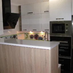 Jasna kuchnia z białymi szafkami, czarnym okapem i blatem w kolorze jasnego drewna, dekoracyjna listwa z motywem owoców nad blatem, zabudowany piekarnik i mikrofalówka.