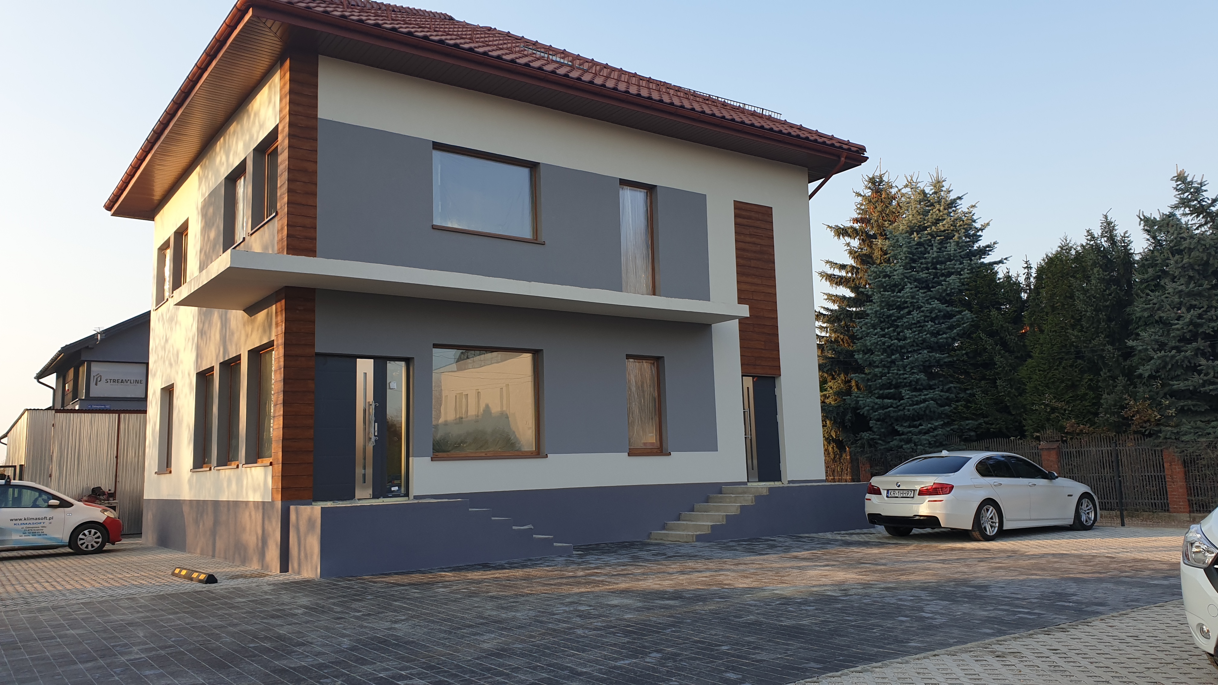 Nowoczesny, dwupiętrowy dom w Krakowie z szarymi i drewnianymi akcentami na elewacji, widoczny z poziomu brukowanego podjazdu, z zaparkowanymi samochodami.