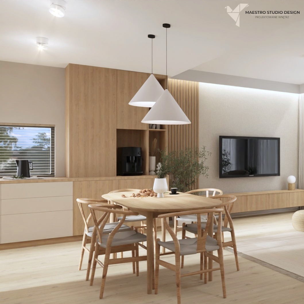 Jasne wnętrze jadalni z drewnianym stołem i krzesłami, minimalistyczne lampy wiszące, zabudowa kuchenna z drewnianymi frontami i czarnym ekspresem do kawy, telewizor na ścianie w tle.
