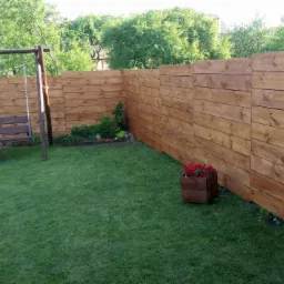 Świeżo zaimpregnowane drewniane ogrodzenie panelowe z huśtawką w tle, otoczone zielonym trawnikiem i roślinami, drewniana donica z czerwonymi kwiatami.