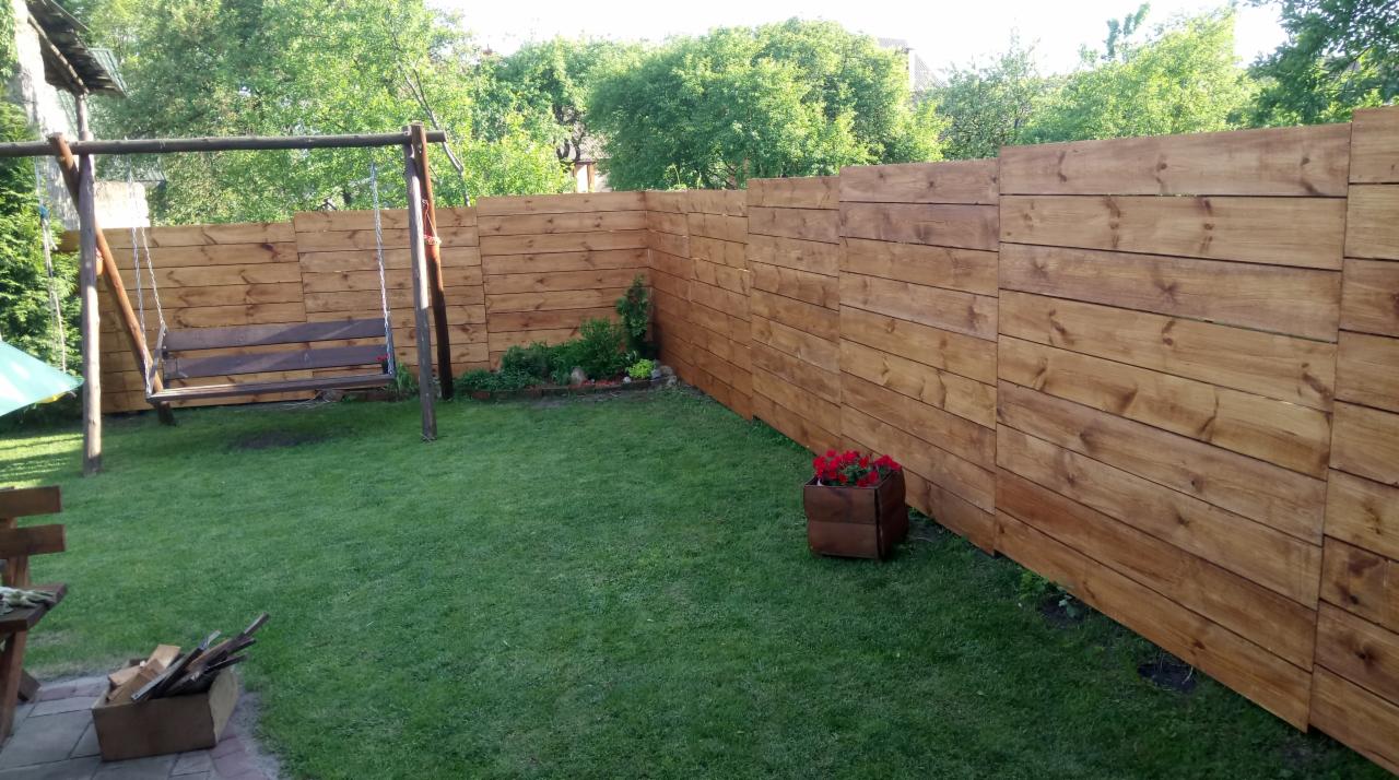 Świeżo zaimpregnowane drewniane ogrodzenie panelowe z huśtawką w tle, otoczone zielonym trawnikiem i roślinami, drewniana donica z czerwonymi kwiatami.