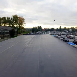Widok z góry na rozległe, płaskie pokrycie dachowe z szarej membrany PVC, sąsiadujące z parkingiem pełnym samochodów i budynkiem w tle, sfotografowane w pochmurny dzień.