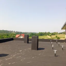 Czarna membrana na płaskim dachu z widocznymi kominami wentylacyjnymi, mocowaniami i fragmentem budynku w trakcie budowy, w tle krajobraz z zabudowaniami i zielenią.