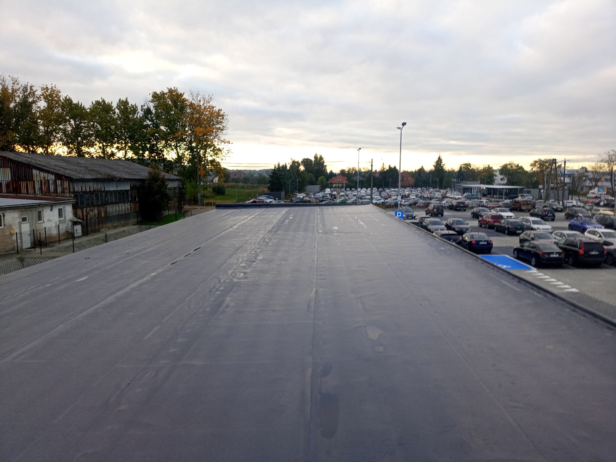 Widok z góry na rozległe, płaskie pokrycie dachowe z szarej membrany PVC, sąsiadujące z parkingiem pełnym samochodów i budynkiem w tle, sfotografowane w pochmurny dzień.