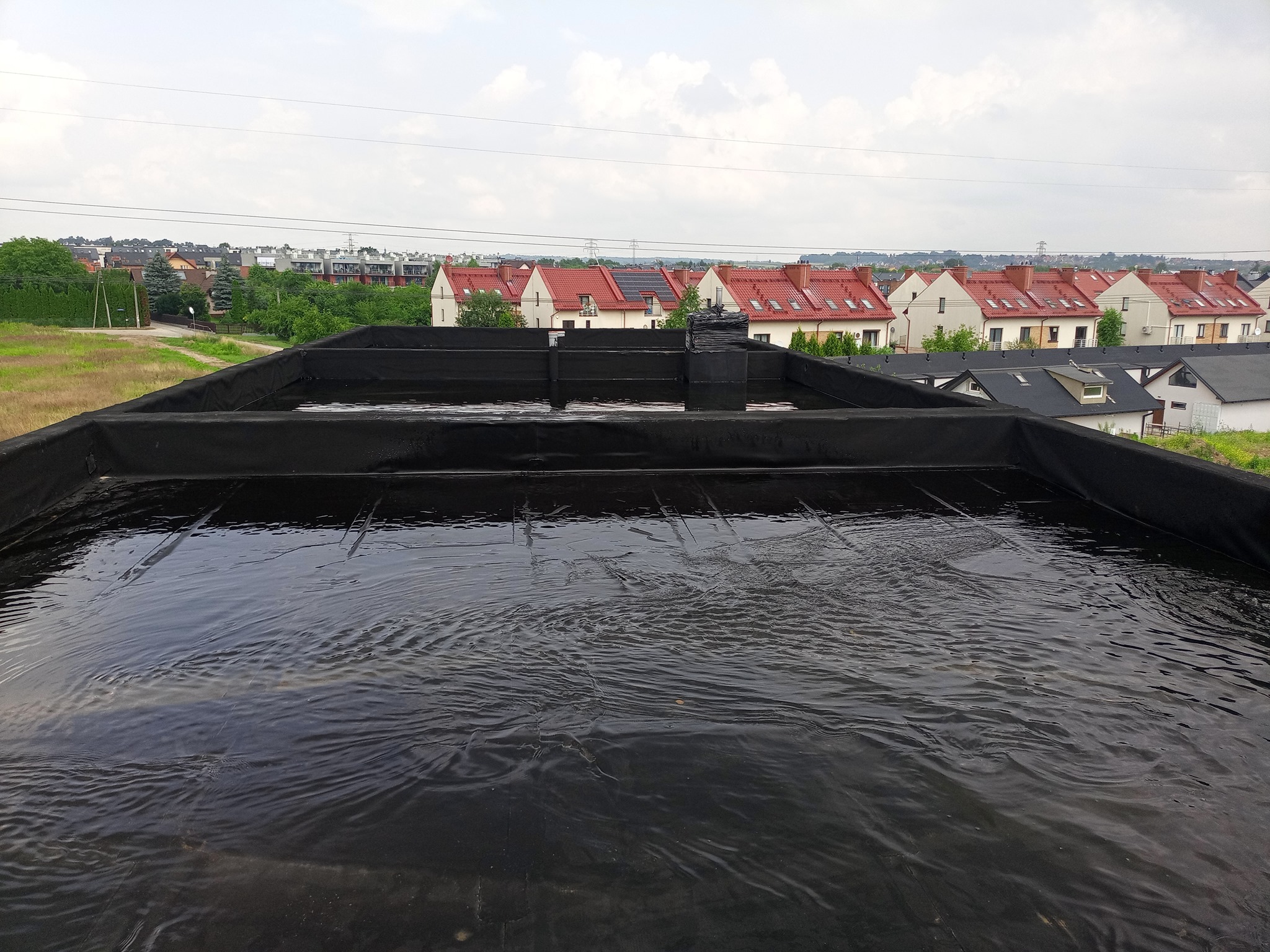 Czarna, płaska powierzchnia dachu pokryta wodą po deszczu, w tle widok na osiedle domów jednorodzinnych i pochmurne niebo.