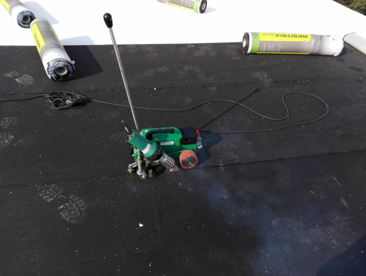 Proces zgrzewania papy termozgrzewalnej na płaskim dachu, widoczne rolki papy, zgrzewarka automatyczna i ślady butów na powierzchni dachu.