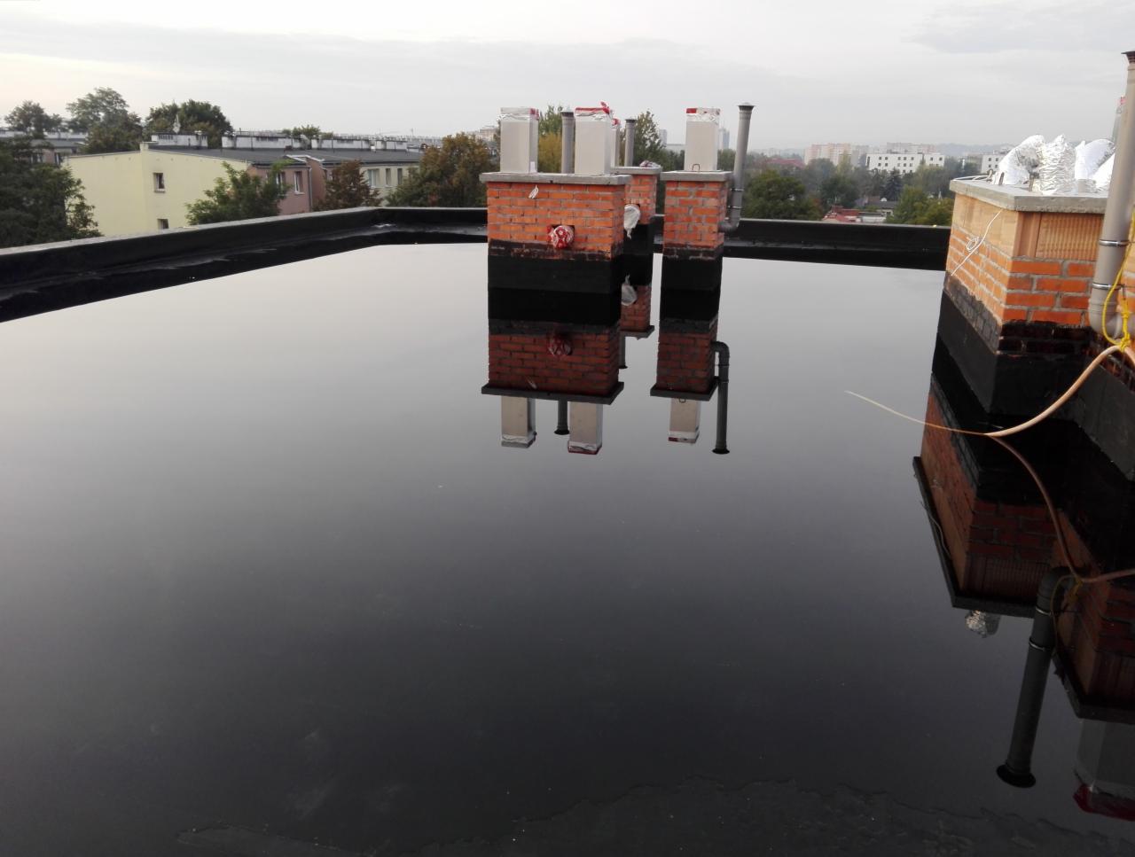 Płaski dach pokryty czarną membraną bitumiczną z odbijającymi się w kałużach kominami wentylacyjnymi, widok na miasto w tle.