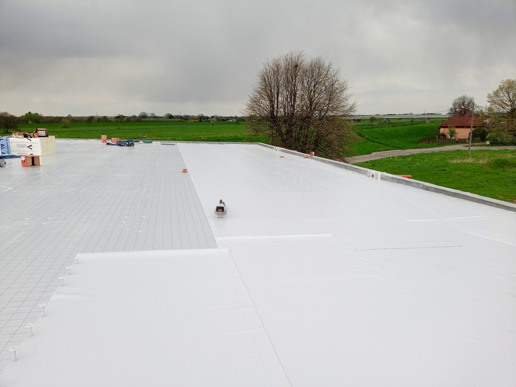 Biała membrana dachowa rozkładana na płaskim dachu, widoczne narzędzia i materiały budowlane, w tle zielony krajobraz i pochmurne niebo.