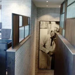 Wnętrze restauracji z ceglaną ścianą, drewnianymi elementami oddzielającymi stoliki i dużym czarno-białym portretem kucharza w tle.