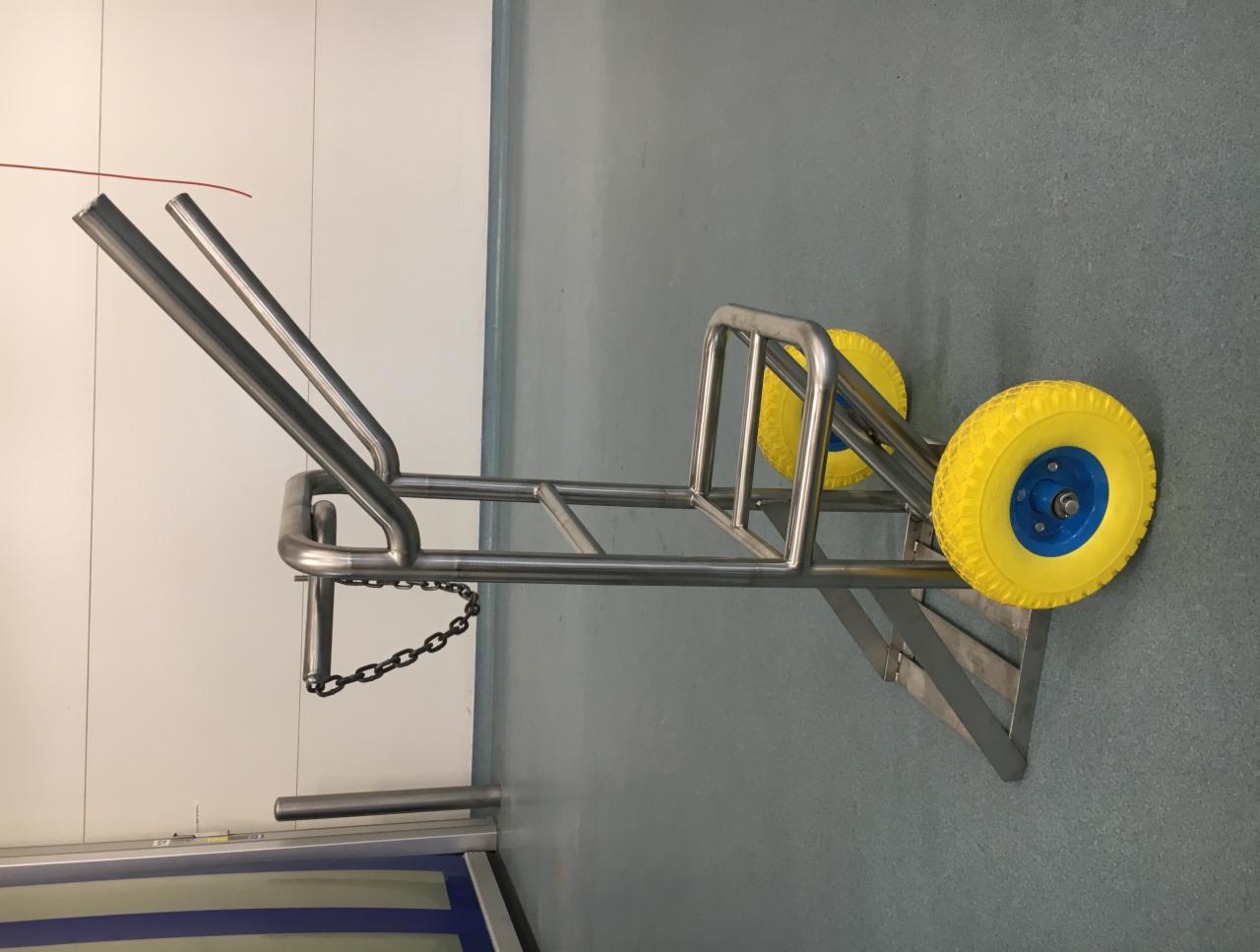 Wózek transportowy ze stali nierdzewnej z żółtymi kołami na niebieskich piastach, łańcuch zabezpieczający zwisa po boku, widok z boku na tle białej ściany i szarej podłogi.