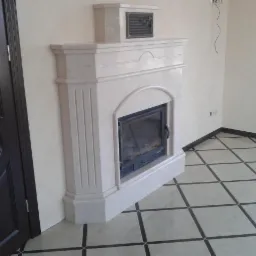 Wykończony narożny kominek z jasnego marmuru z czarnym wkładem kominkowym, na podłodze jasne płytki w ciemną kratę, widok z boku drzwi.