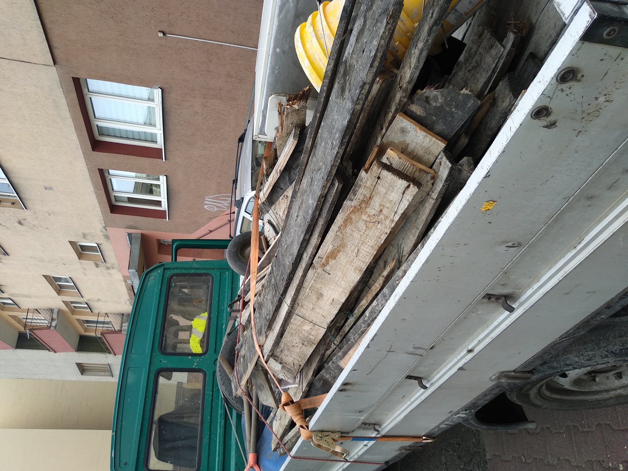 Załadunek drewnianych desek i elementów konstrukcyjnych na skrzynię samochodu ciężarowego, zabezpieczonych pomarańczowymi pasami transportowymi, w tle zabudowa miejska.