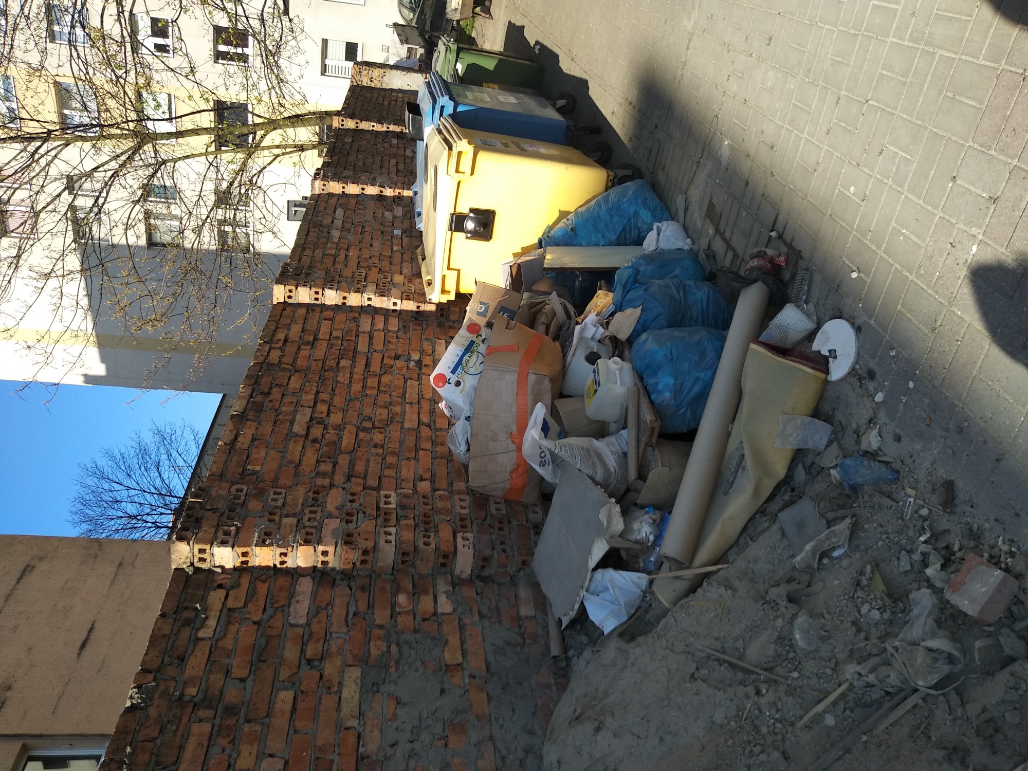 Sterta odpadów budowlanych i gruzu obok śmietników, przy ceglanym murze i chodniku z kostki, w tle budynek mieszkalny i drzewo.