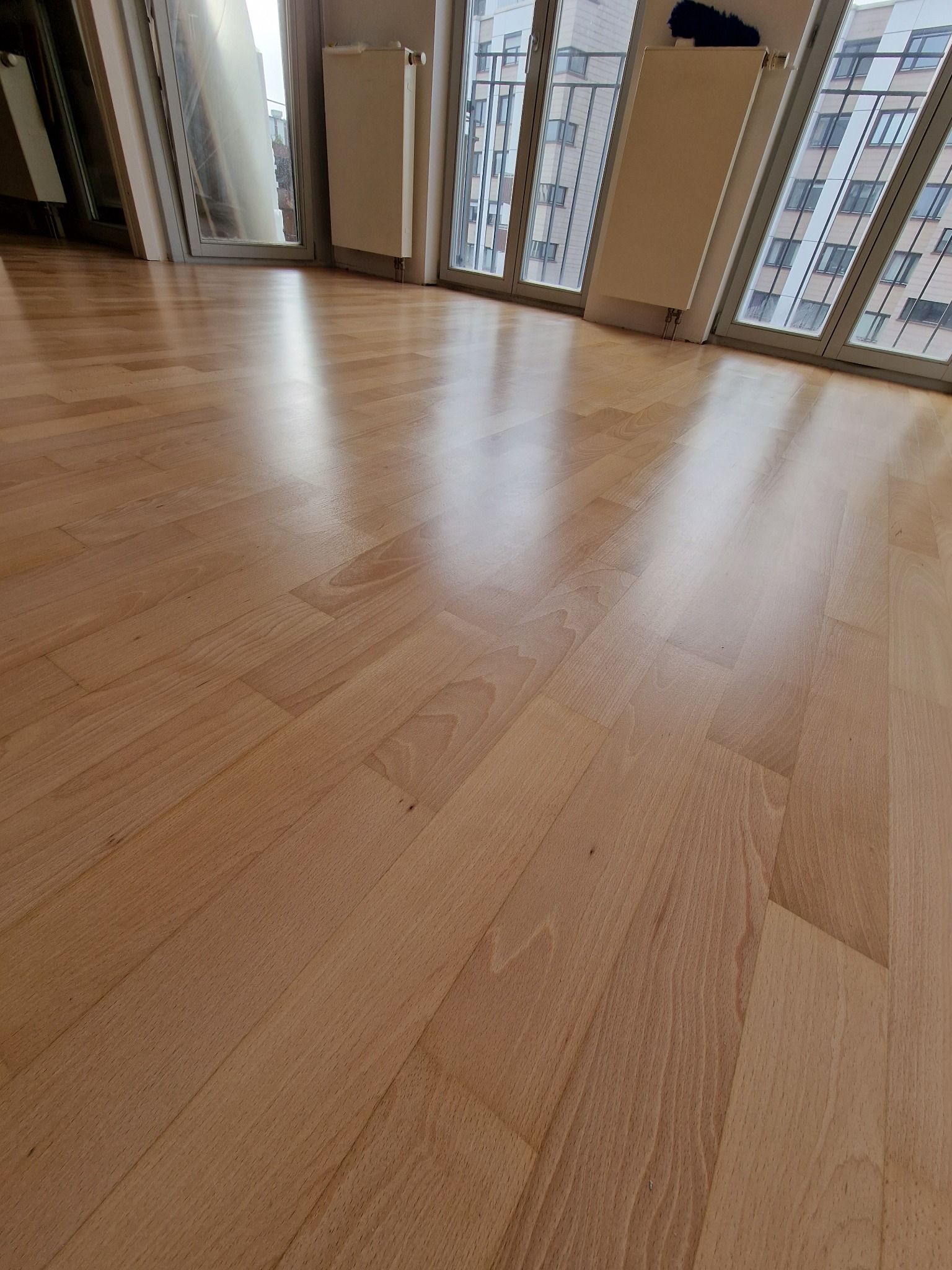 Jasny, lakierowany parkiet ułożony w nowym mieszkaniu z dużymi oknami i grzejnikami.