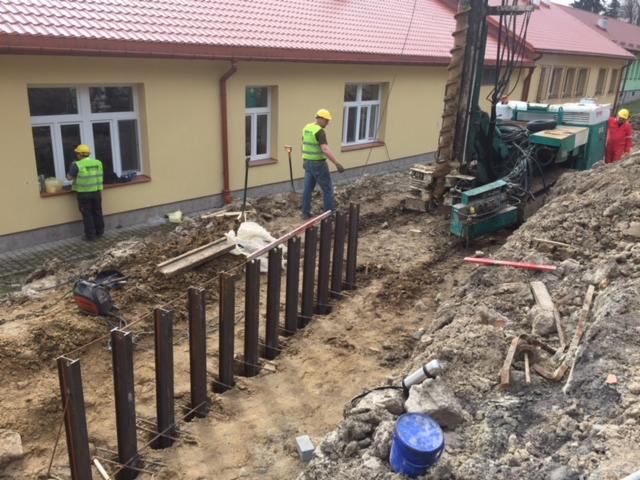 Wbijanie ścianki szczelnej z profili stalowych w wykopie, widoczna wiertnica, robotnicy w kamizelkach odblaskowych i kaskach, budynek w tle.