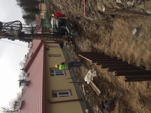 Budowa drogi: robotnicy w kaskach i kamizelkach odblaskowych pracują przy wzmacnianiu skarpy przy użyciu stalowych elementów, z tyłu widoczny budynek z czerwoną dachówką i maszyna budowlana.