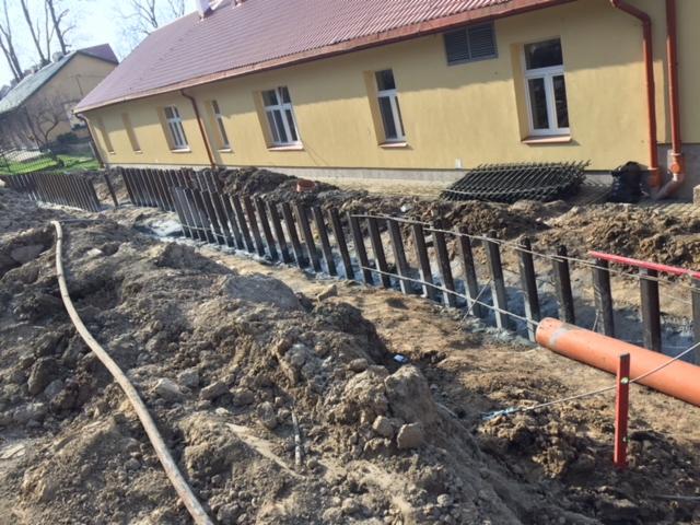 Wykop z zabezpieczeniem ścianek pionowymi belkami stalowymi, widoczny pomarańczowy rurociąg i żółty budynek w tle.