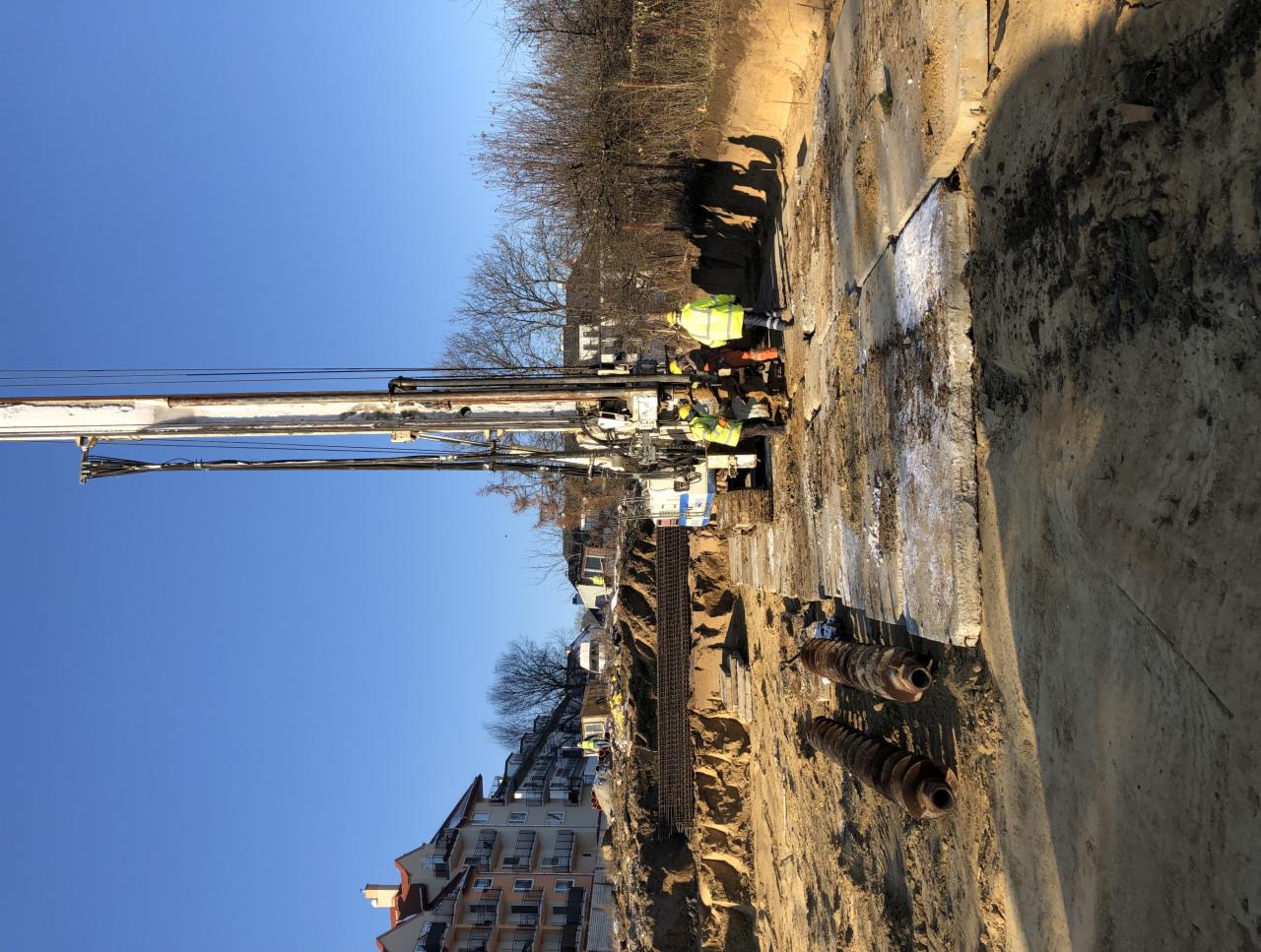 Prace ziemne przy użyciu wiertnicy na placu budowy, widoczni pracownicy w odblaskowych kamizelkach, przygotowanie terenu pod fundamenty.