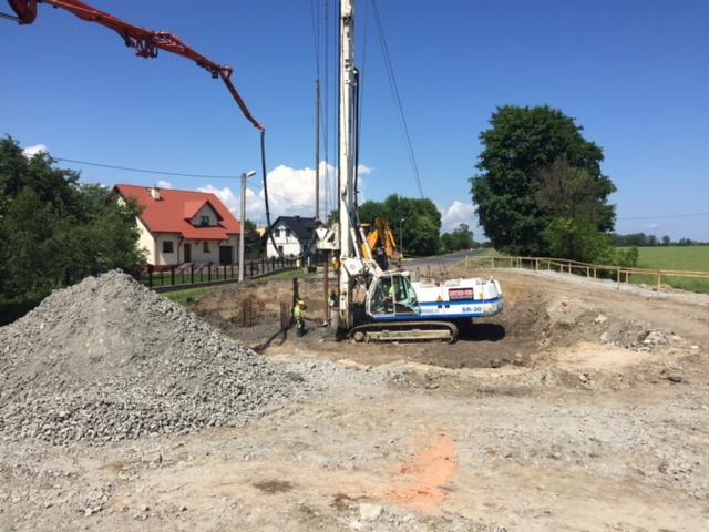 Biała wiertnica SR-30 pracuje przy budowie drogi, w tle widoczny dom z czerwonym dachem i sterta gruzu.