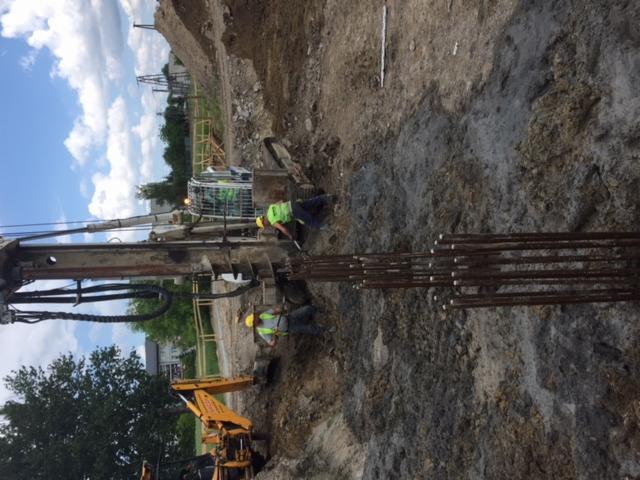 Dwóch robotników w żółtych kamizelkach przy wiertnicy, przygotowujących zbrojenie w wykopie pod fundamenty