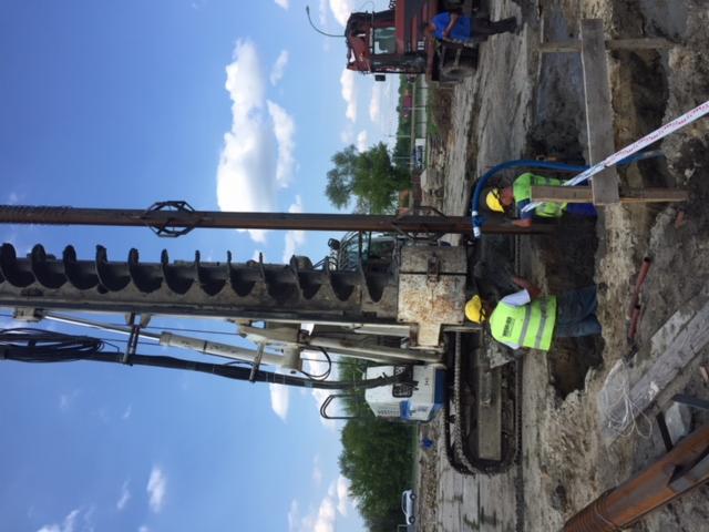 Prace ziemne przy użyciu wiertnicy, dwóch pracowników w żółtych kaskach i kamizelkach odblaskowych, zabezpieczenie taśmą ostrzegawczą, widoczny fragment nieba i drzew w tle.