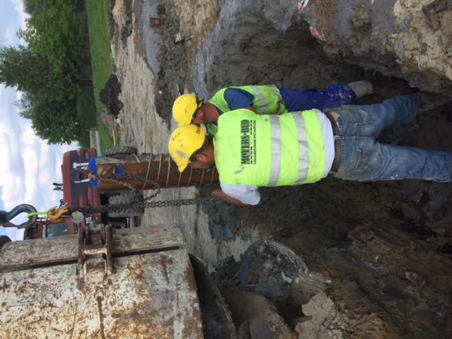 Dwóch pracowników w żółtych kaskach i kamizelkach odblaskowych pracuje w wykopie przy użyciu ciężkiego sprzętu, prawdopodobnie układając rury.