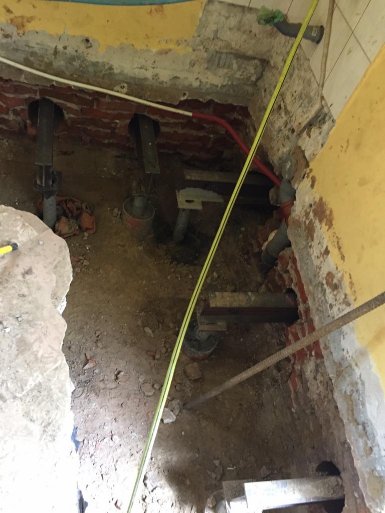 Wykop z odsłoniętymi fundamentami budynku, wzmocnionymi stalowymi podporami i widocznymi instalacjami, z fragmentem żółtej miarki zwisającej wzdłuż ściany.