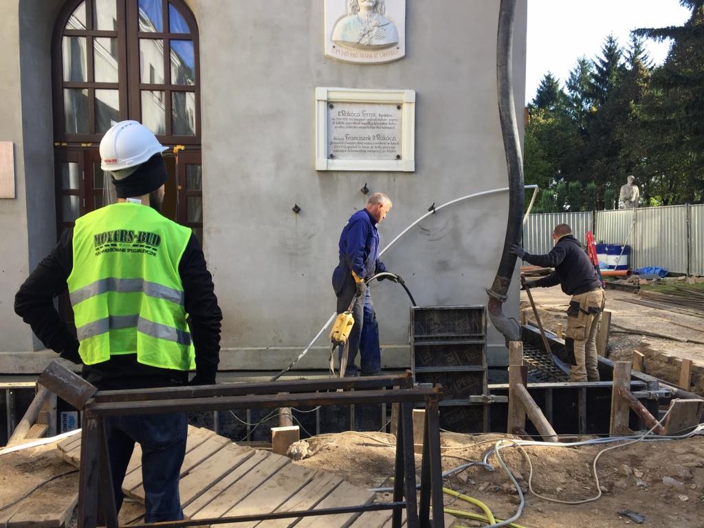 Pracownicy w trakcie wylewania fundamentów pod budynek z elementami architektonicznymi, widoczny szalunek, pompa do betonu i pracownik w kamizelce odblaskowej z napisem Moxers-Bud.