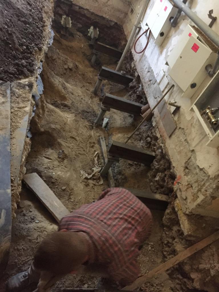 Pracownik w koszuli w kratę w wykopie ziemnym zabezpieczonym drewnianymi schodami i podporami, widoczne instalacje na ścianie budynku.