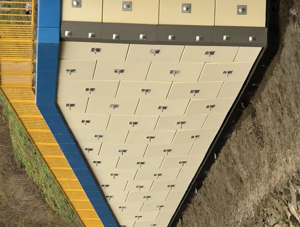 Fragment konstrukcji oporowej z paneli betonowych, zabezpieczającej skarpę przy drodze z żółtą barierką.