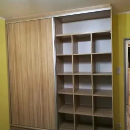 Szafa przesuwna z drewnopodobnymi frontami i otwartymi półkami, widoczna na tle żółtej ściany i drewnianej podłogi.