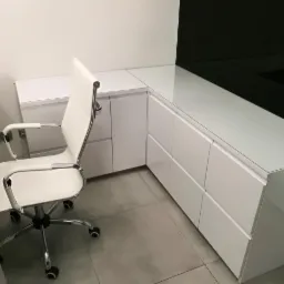 Białe biurko z szufladami i czarnym panelem ściennym, uzupełnione o białe krzesło biurowe na kółkach.