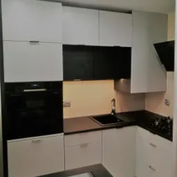 Nowoczesna kuchnia z białymi frontami i czarnymi akcentami w Luszowicach, widok na zabudowę kuchenną z piekarnikiem, płytą grzewczą i zlewozmywakiem.