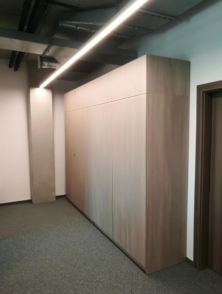 Duża, jasna szafa wnękowa z drewnopodobnym wykończeniem, zamontowana w biurze z szarym dywanem i widocznymi elementami instalacji pod sufitem.
