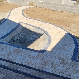 ŻUK-BRUK - Ścieżka z kostki brukowej w odcieniach beżu i szarości, wijąca się przez teren budowy. Widoczny fragment fundamentów budynku w tle. Ujęcie z góry.