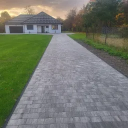Szeroki podjazd z szarej kostki brukowej prowadzący do nowoczesnego domu z garażem, oświetlony ciepłym światłem zachodzącego słońca.
