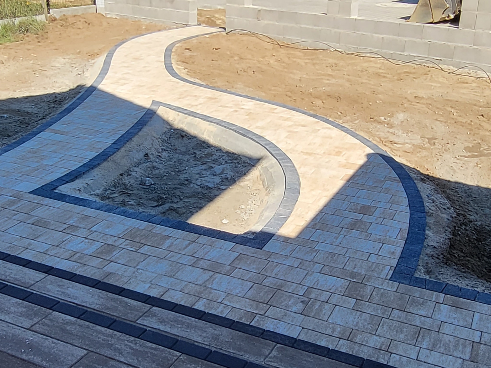 Ścieżka z kostki brukowej w odcieniach beżu i szarości, wijąca się przez teren budowy. Widoczny fragment fundamentów budynku w tle. Ujęcie z góry.