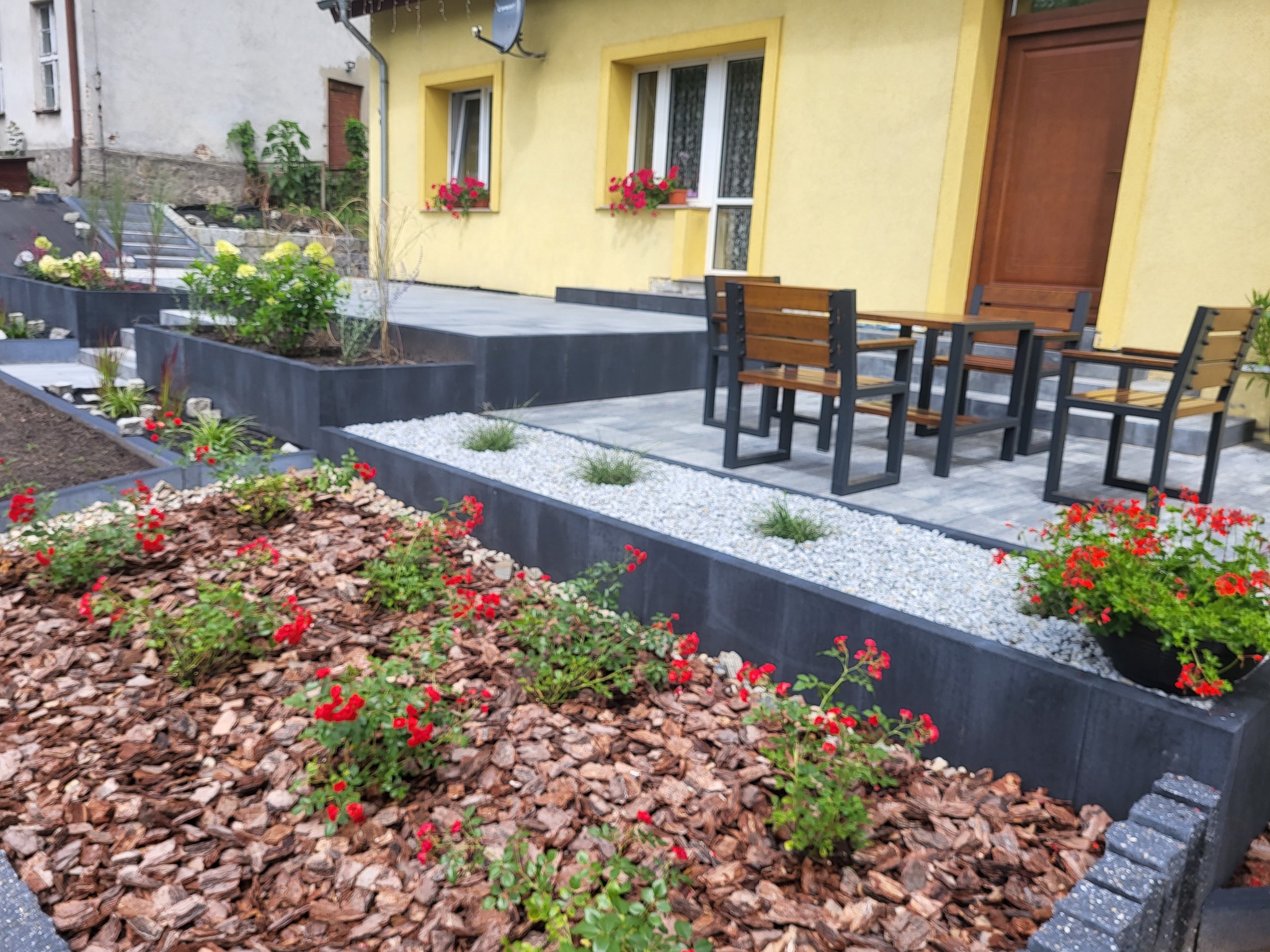 Nowoczesny ogród z podwyższonymi rabatami z kostki, wysypany korą i żwirem, z kwitnącymi czerwonymi różami i zestawem mebli ogrodowych na tle żółtego domu.